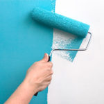 A hand with a paint roller covered in teal blue paint, painting over a white wall