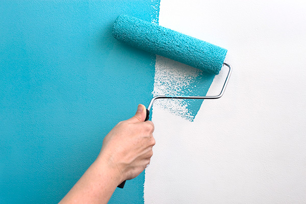 A hand with a paint roller covered in teal blue paint, painting over a white wall