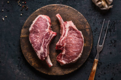 Marbled raw pork chops of Porco Iberico meat on round cutting board with meat knife on dark rustic background, top view. French Racks
