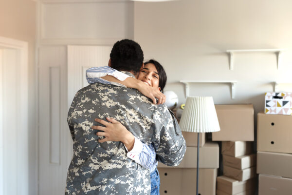 Wife embraces her army veteran husband in camouflage clothing while they moving into the new house. First time home buyers celebrate and hugging new house purchase on moving day, laughing standing among boxes in own flat house