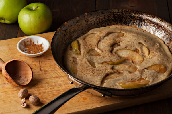 A high angle extreme close up horizontal photograph of a German apple pancake in grandmas well used frying pan. A bowl with ground cinnamon and a small wooden scoop along with ground nutmeg and some whole nutmegs sit by the pan on the cutting board.Two fresh green granny smith apple can be seen in the background.
