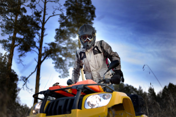 Horizontal close-up of a man in helmet and safety goggles looking into the camera while sitting on quad bike against vivid blue sky.