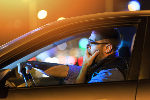 Side view of young man with eyeglasses sitting inside of his car and yawning. One hand on mouth. Bright lights at background.