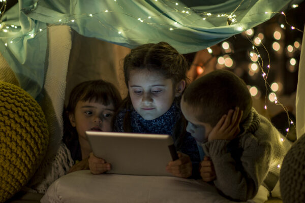 Two Caucasian girls and a Caucasian boy are indoors. They are siblings. They are inside a pillow house decorated with glowing lights. They are wearing casual clothing. They are watching a movie on a tablet computer.