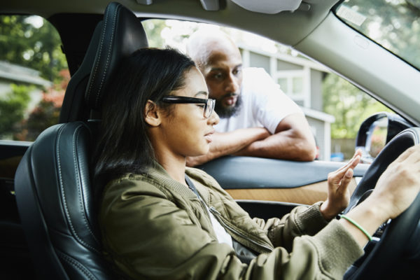 Father leaning through window of car while teaching daughter to drive